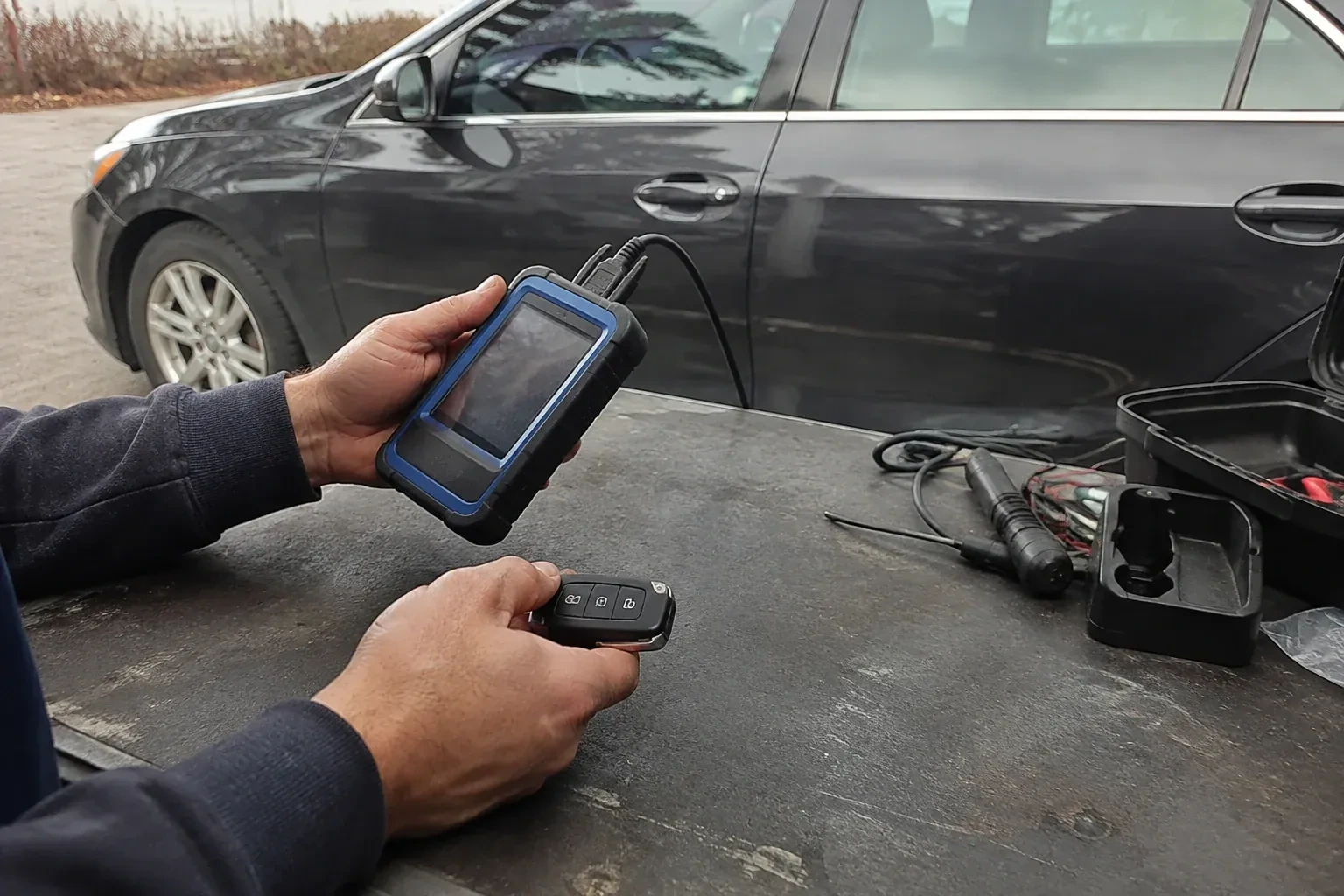 Hands holding a blue-edged electronic device and car-key fob at a worktable beside a parked car. — Transponder Key Programming, Guelph