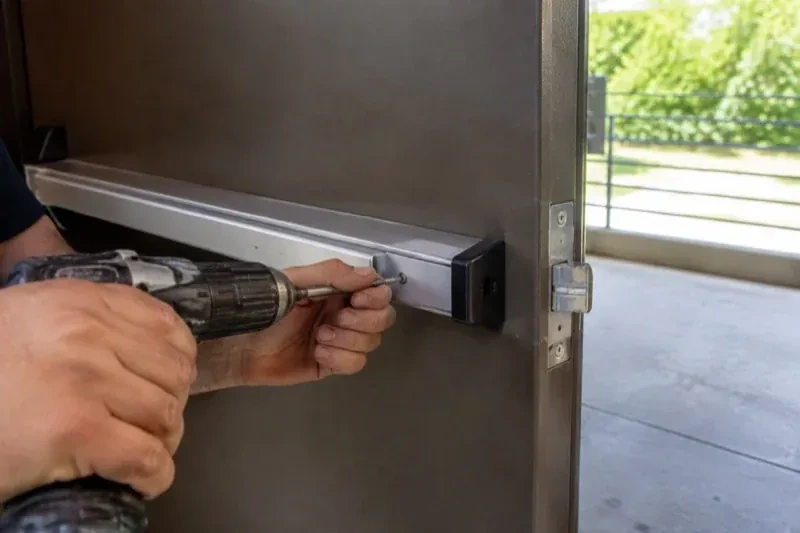 Hands using a power driver at the end of a horizontal push bar on an open metal door. — Panic & Exit Hardware, Guelph
